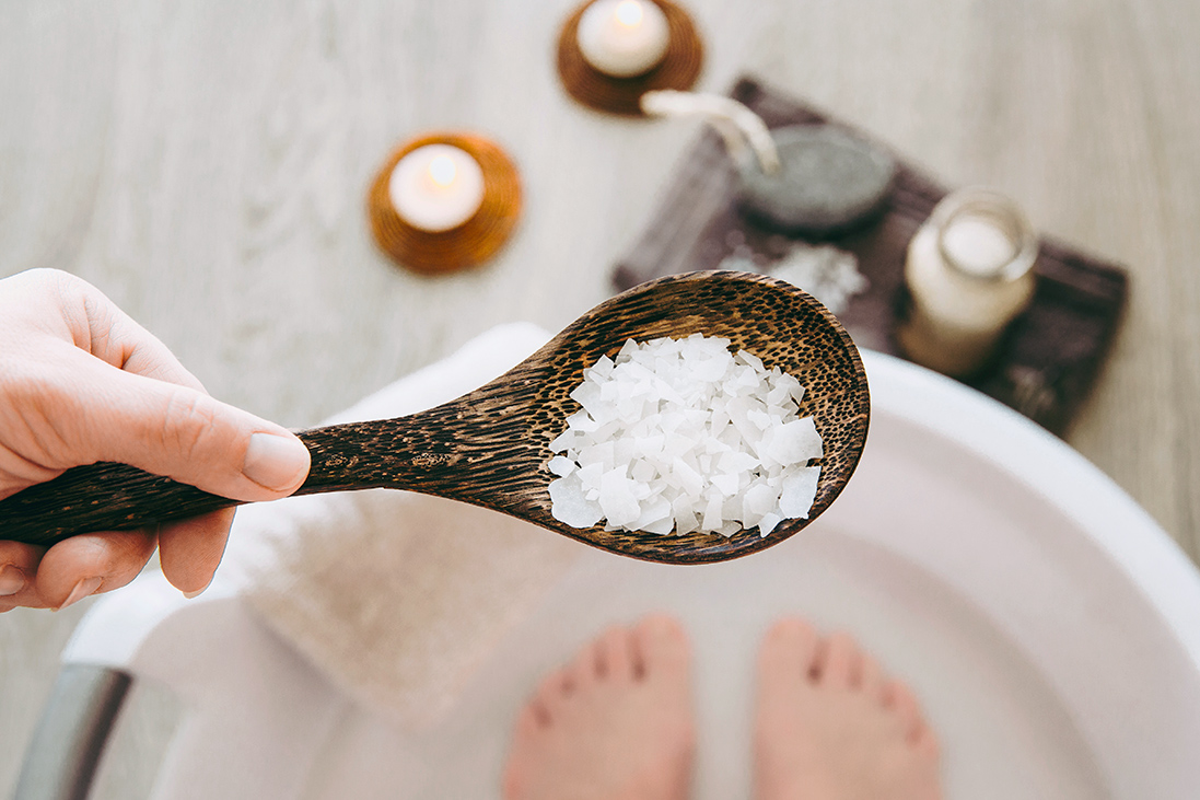 Vamos Ratgeber Hilfe bei Schweißfüßen Hausmittel Bild eine Hand mit Holzlöffel voll Salze gibt diese in eine Badewanne zu den Füßen